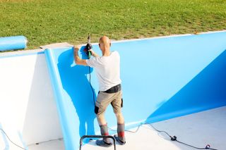 Person spray-painting a bright blue pool wall outdoors with a paint sprayer