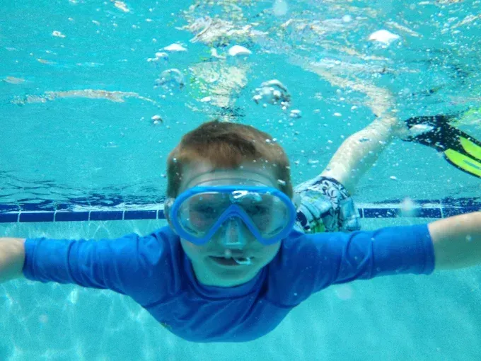 Child swimming underwater in a blue pool, wearing goggles and a blue shirt, arms outstretched.