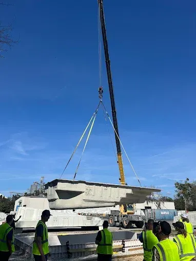 Crane lifting a large white concrete block while workers in neon vests stand nearby under a blue sky