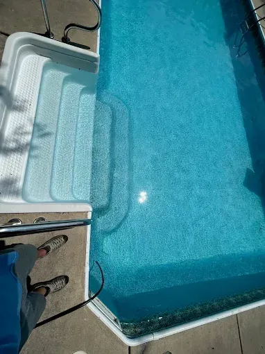 White pool steps beside clear blue water, with a person's shoes visible on the deck.