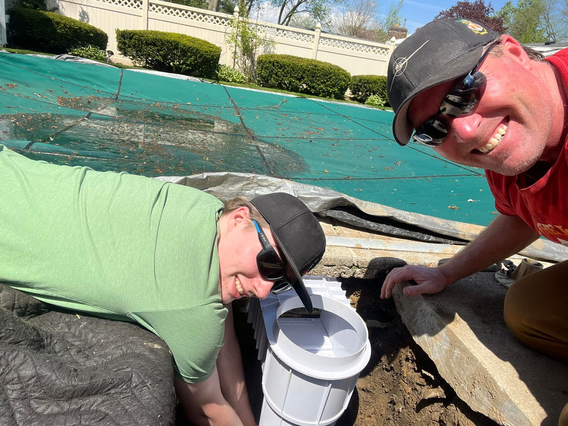 Two people crouch beside a white bucket on a pool deck, with a turquoise pool and fountains behind them.