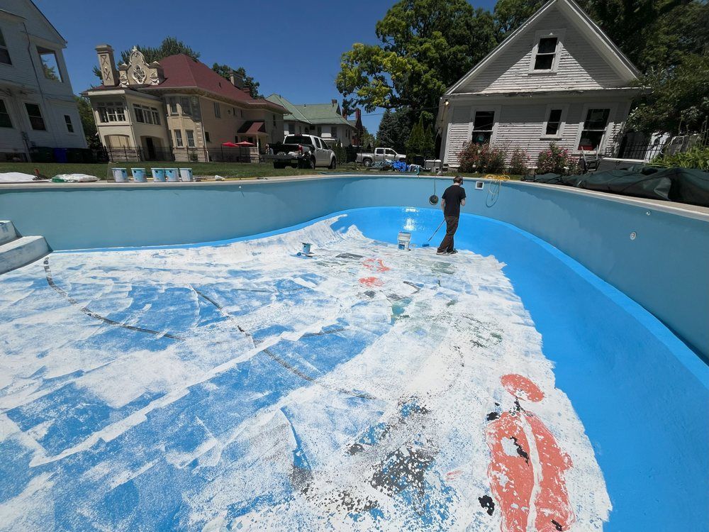 Drained swimming pool being cleaned, with a worker spraying water across the blue liner under a sunny sky