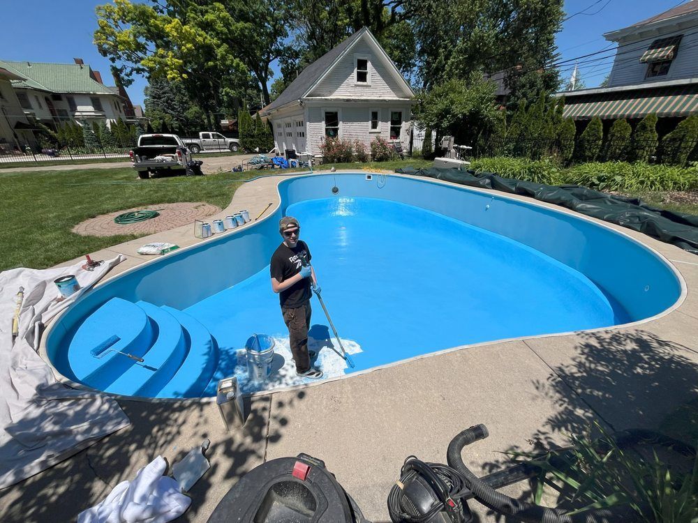 Person standing in a bright blue backyard pool with a net, surrounded by a sunny residential yard.