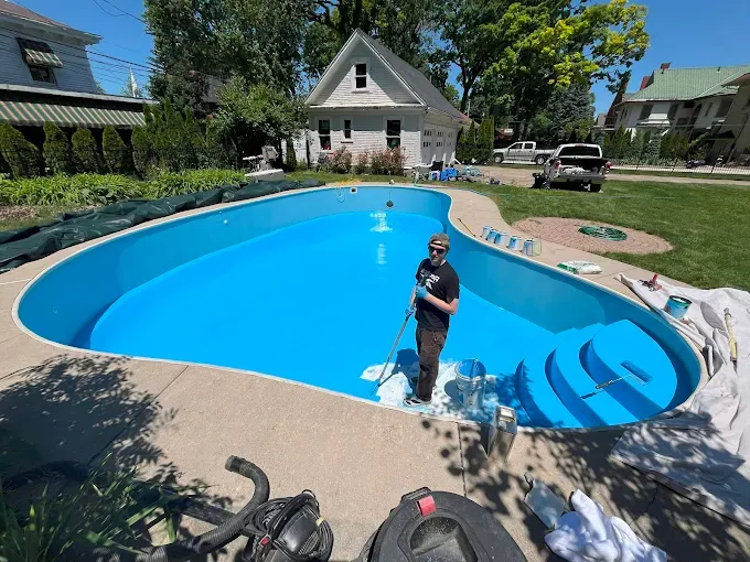 Person standing in a bright blue backyard pool with a diving board and houses in the background