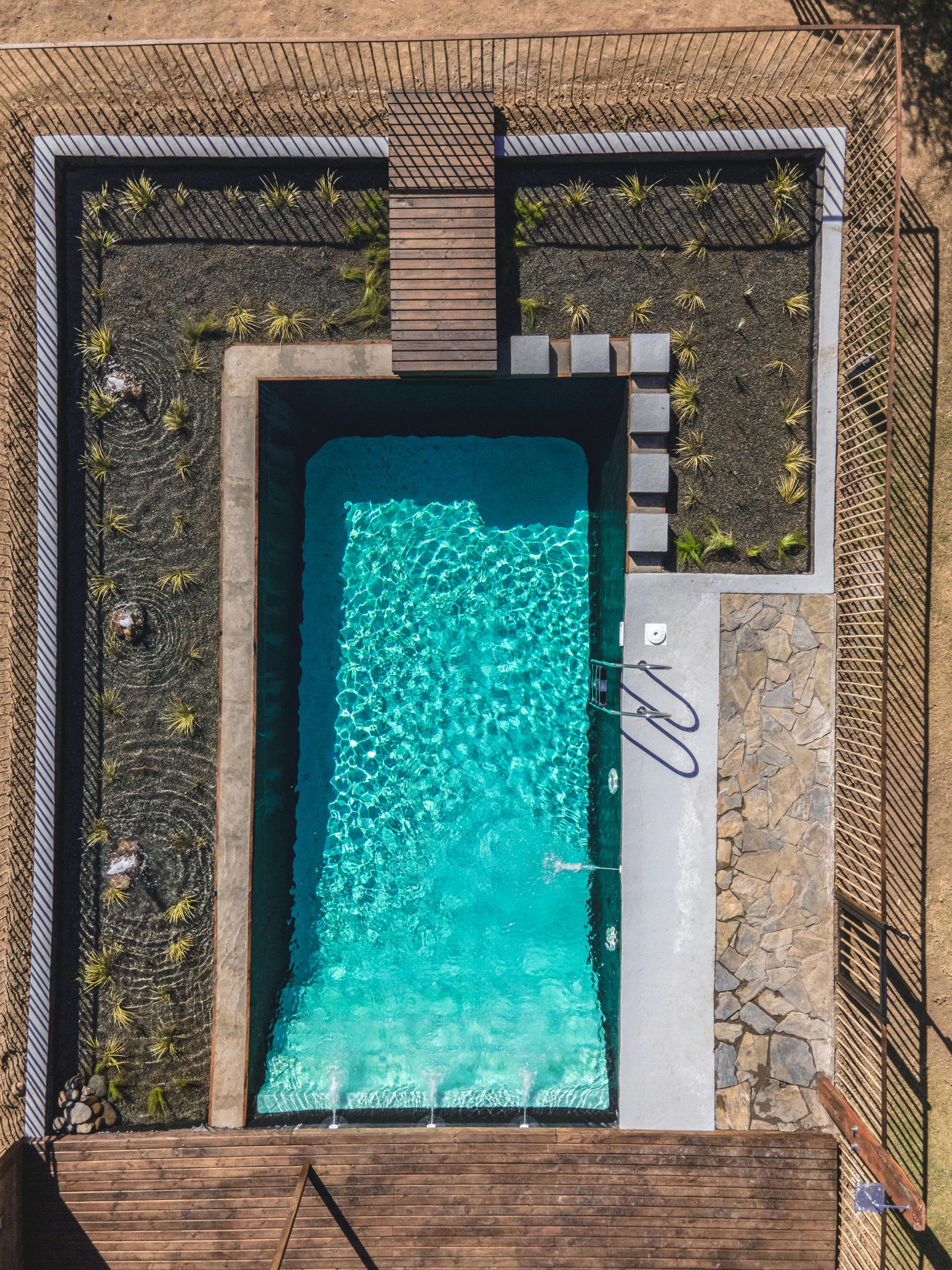 Aerial view of a rectangular turquoise pool beside a white deck and brown stone paving.