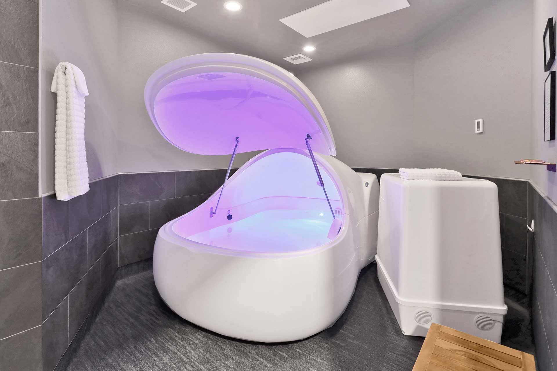 A flotation therapy chamber with purple lighting sits in a tiled room. A towel hangs nearby.