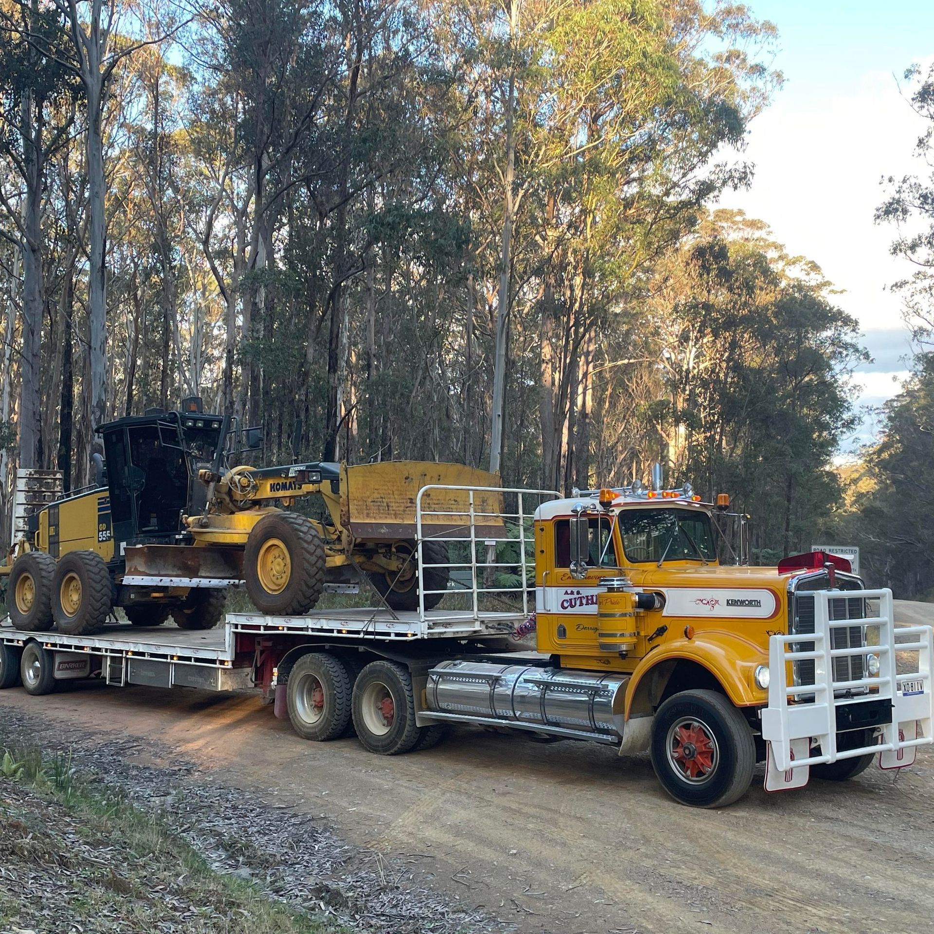Yellow Flatbed Tow Truck Carrying a Heavy Loader on A Forest Road — A & S Cuthel Earthmoving & Transport in Dorrigo, NSW
