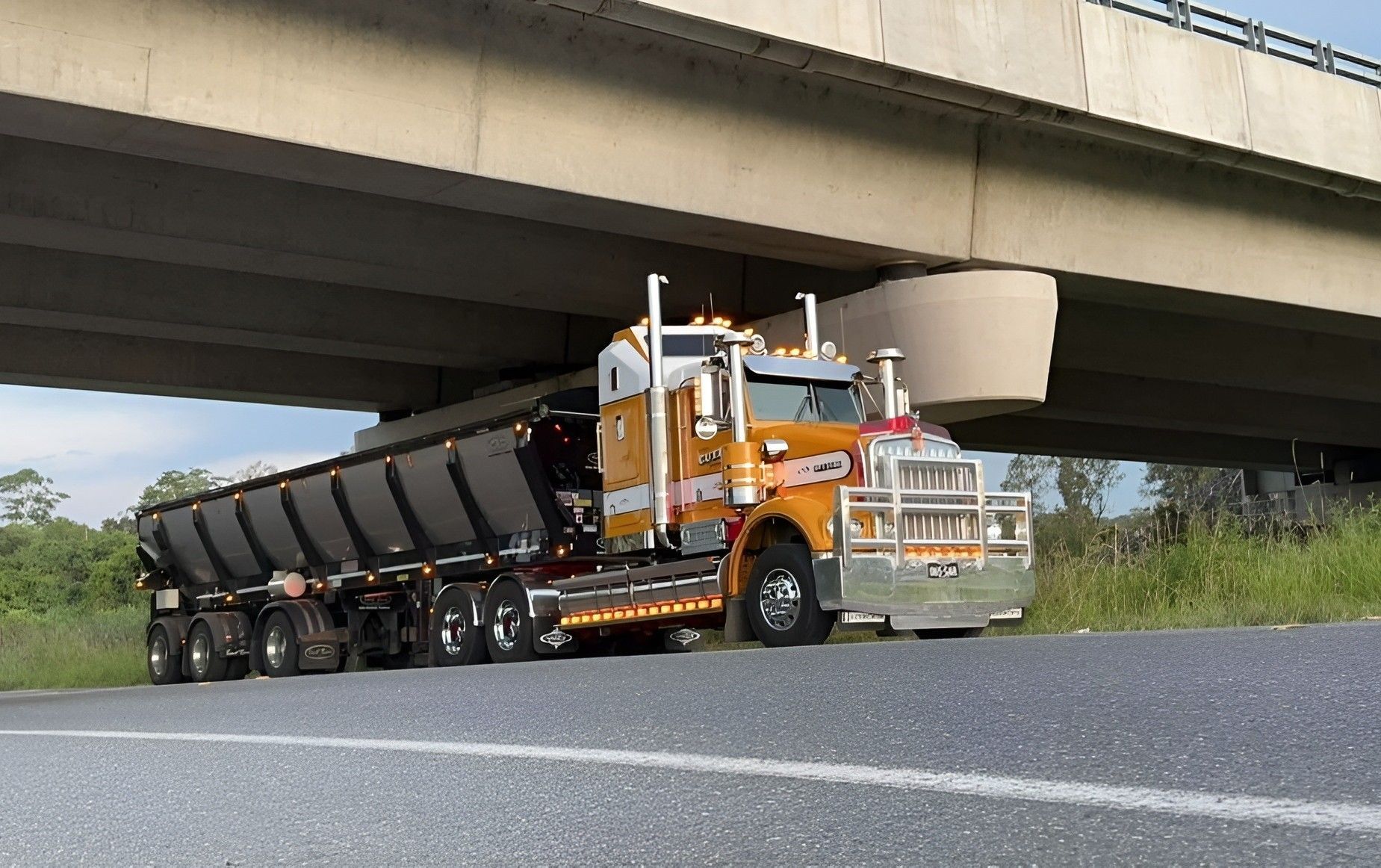 Orange Semi Truck Hauling a Long Black Trailer Under a Concrete Overpass — A & S Cuthel Earthmoving & Transport in Grafton, NSW