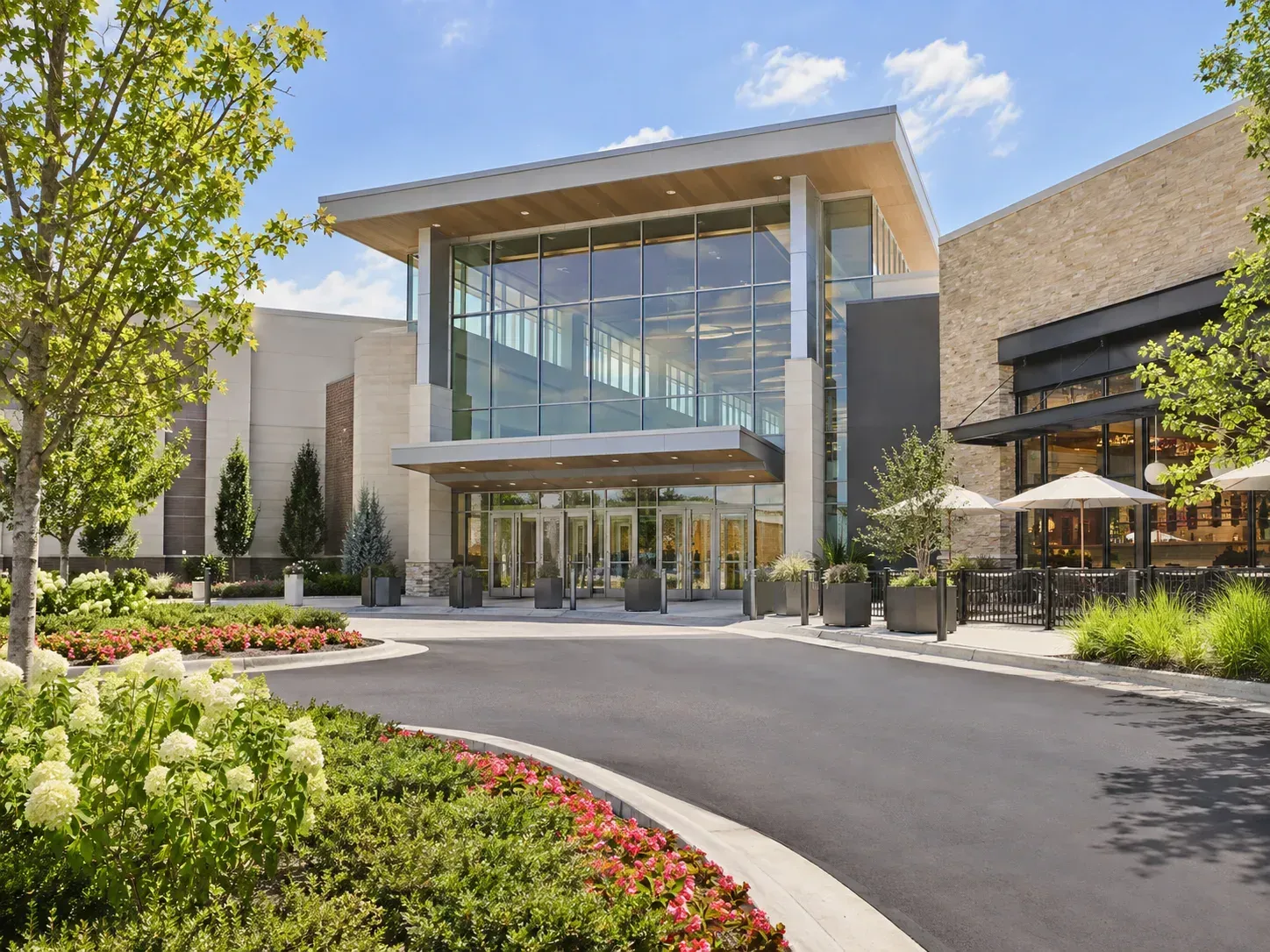 An elevated outdoor view of a modern shopping mall promenade with manicured greenery, palm trees, and pedestrians.