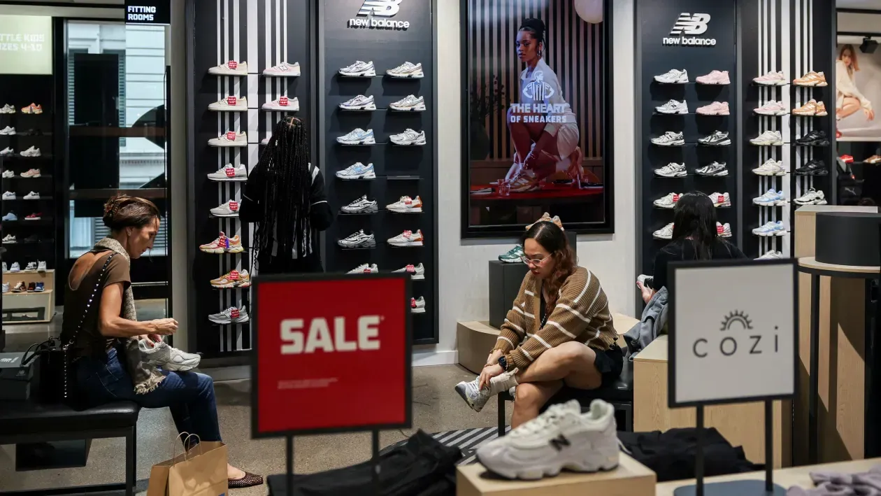 Two people sit in a New Balance store while shopping near a display of shoes and a prominent red sale sign.