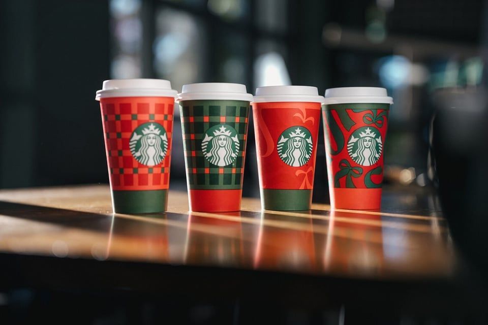 Four Starbucks holiday-themed paper cups with different festive patterns, standing in a row on a wooden table.