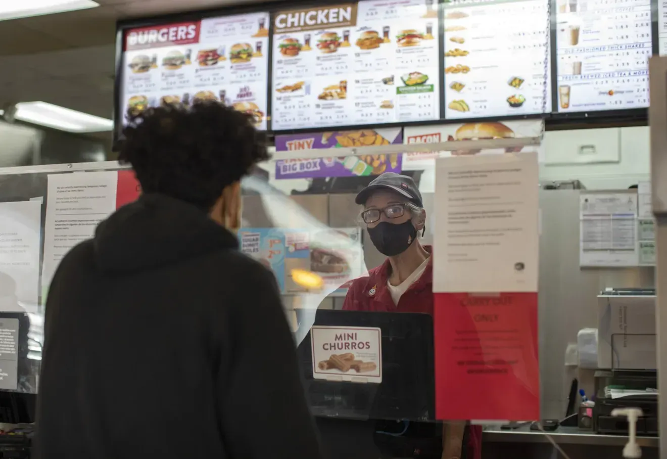 A customer stands at a fast-food counter, facing a masked employee who is preparing to take their order.
