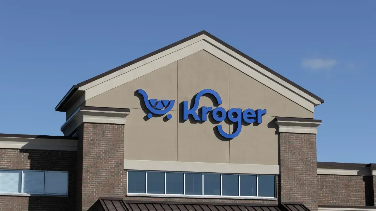 The blue Kroger logo and shopping cart icon mounted on the beige exterior of a brick grocery store under a blue sky.