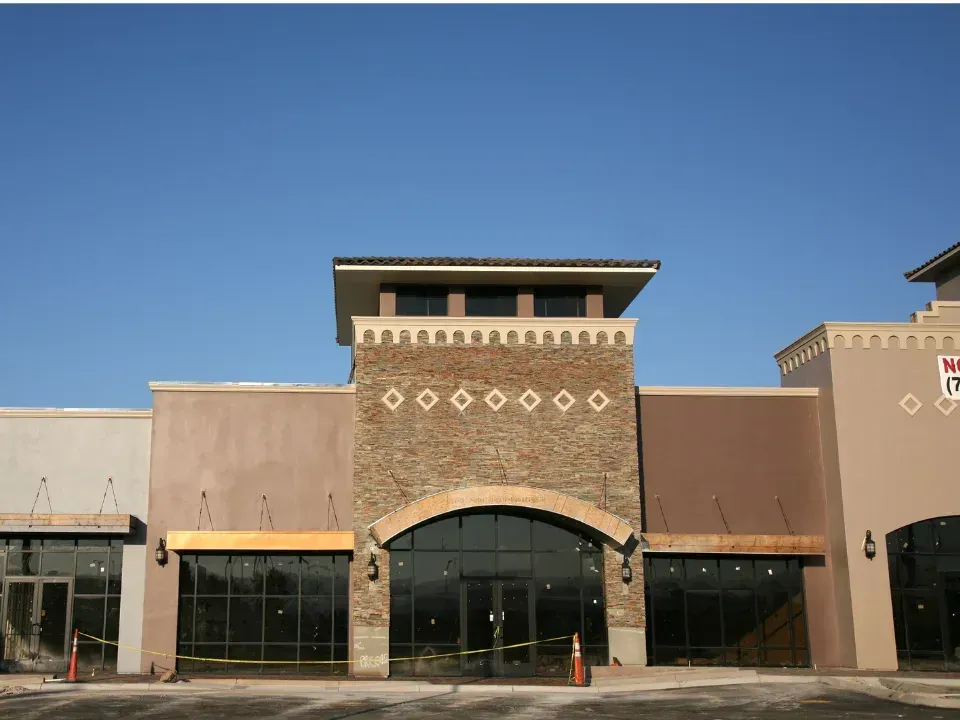 A modern, tan-colored strip mall building under a clear blue sky, featuring a stone-facade entrance with glass storefronts.