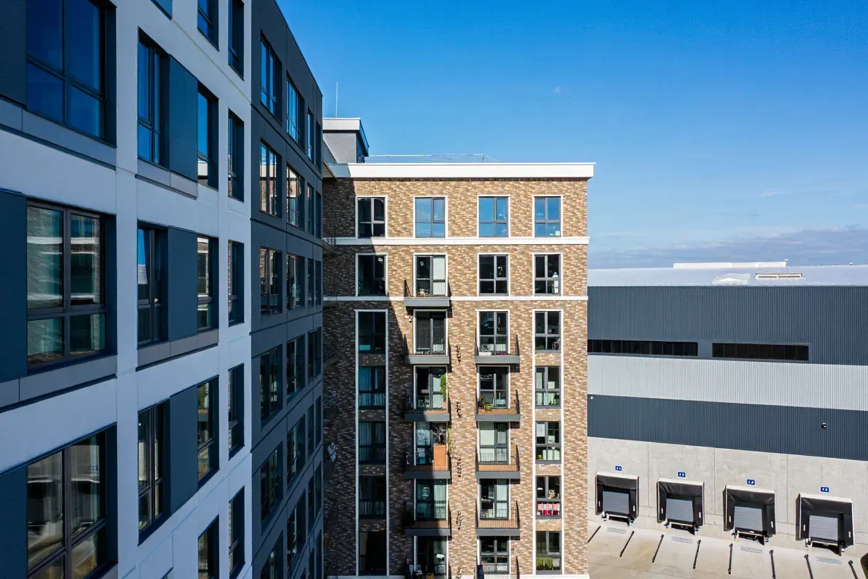 Modern brick apartment building beside a light gray industrial warehouse with loading docks under a clear blue sky.