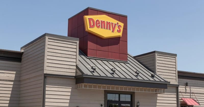 A Denny's restaurant exterior with a yellow sign on a maroon tower, beige horizontal siding, and a dark metal roof.