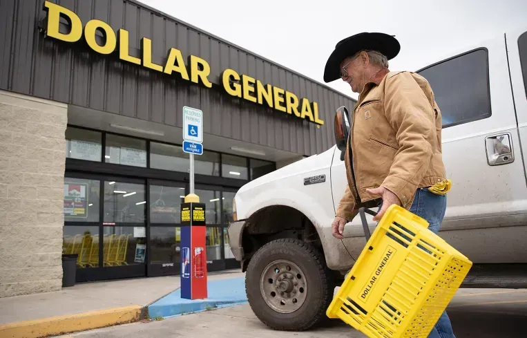 A person wearing a cowboy hat carries a yellow Dollar General basket while walking past a truck at a store entrance.