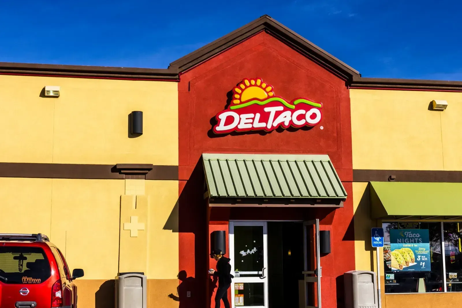 The front entrance of a Del Taco restaurant with a red and yellow facade, a green awning, and a partially visible red car.