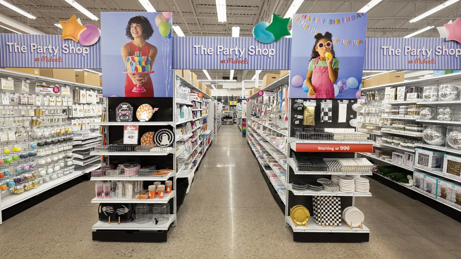 A party supply aisle in a store with colorful signs featuring people holding festive items above stocked shelves.