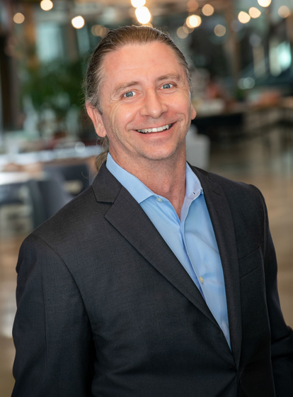 Smiling man in a dark blazer and blue shirt standing in a softly lit indoor setting