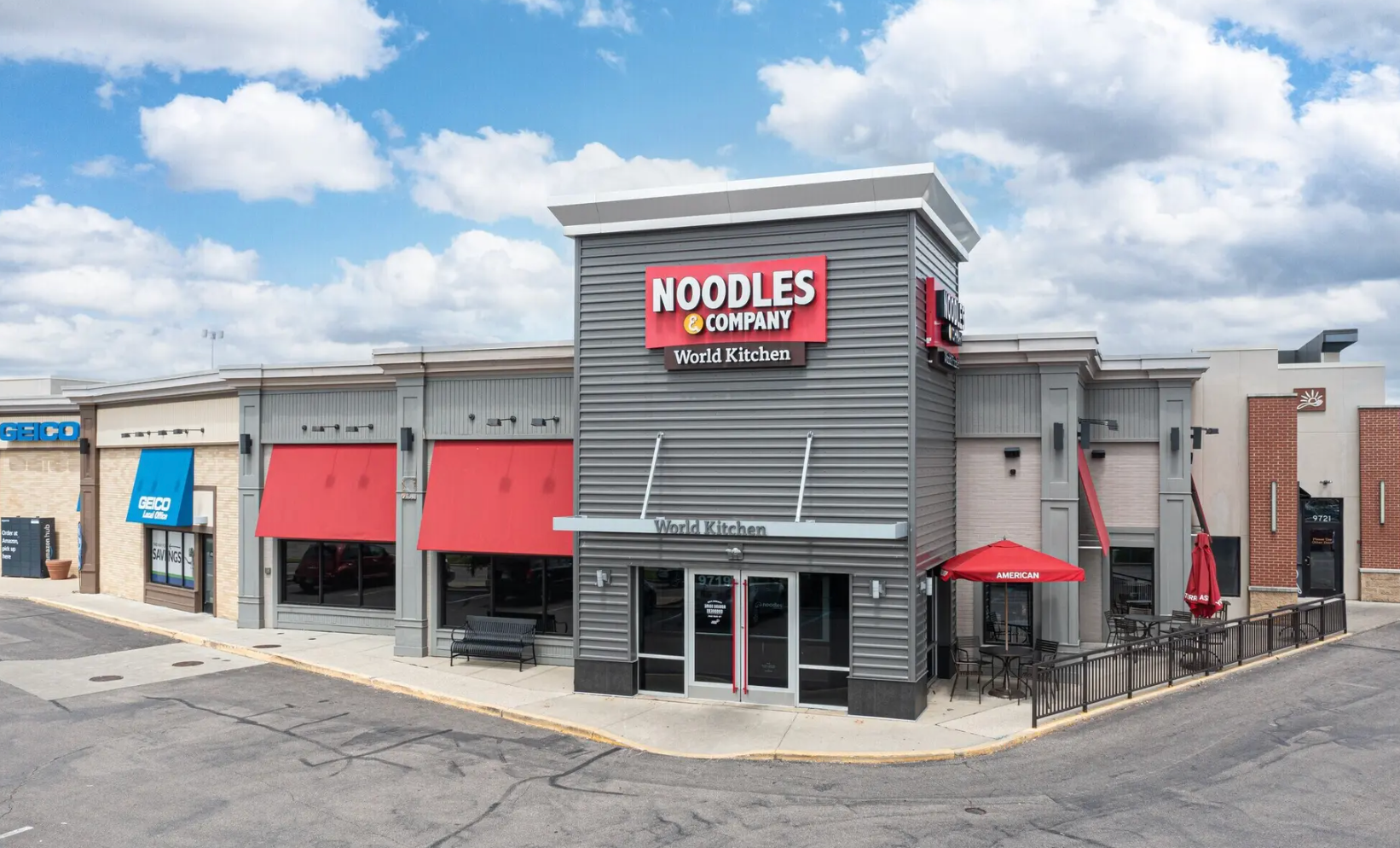 A Noodles & Company restaurant with grey siding, red awnings, and an outdoor patio under a cloudy sky.