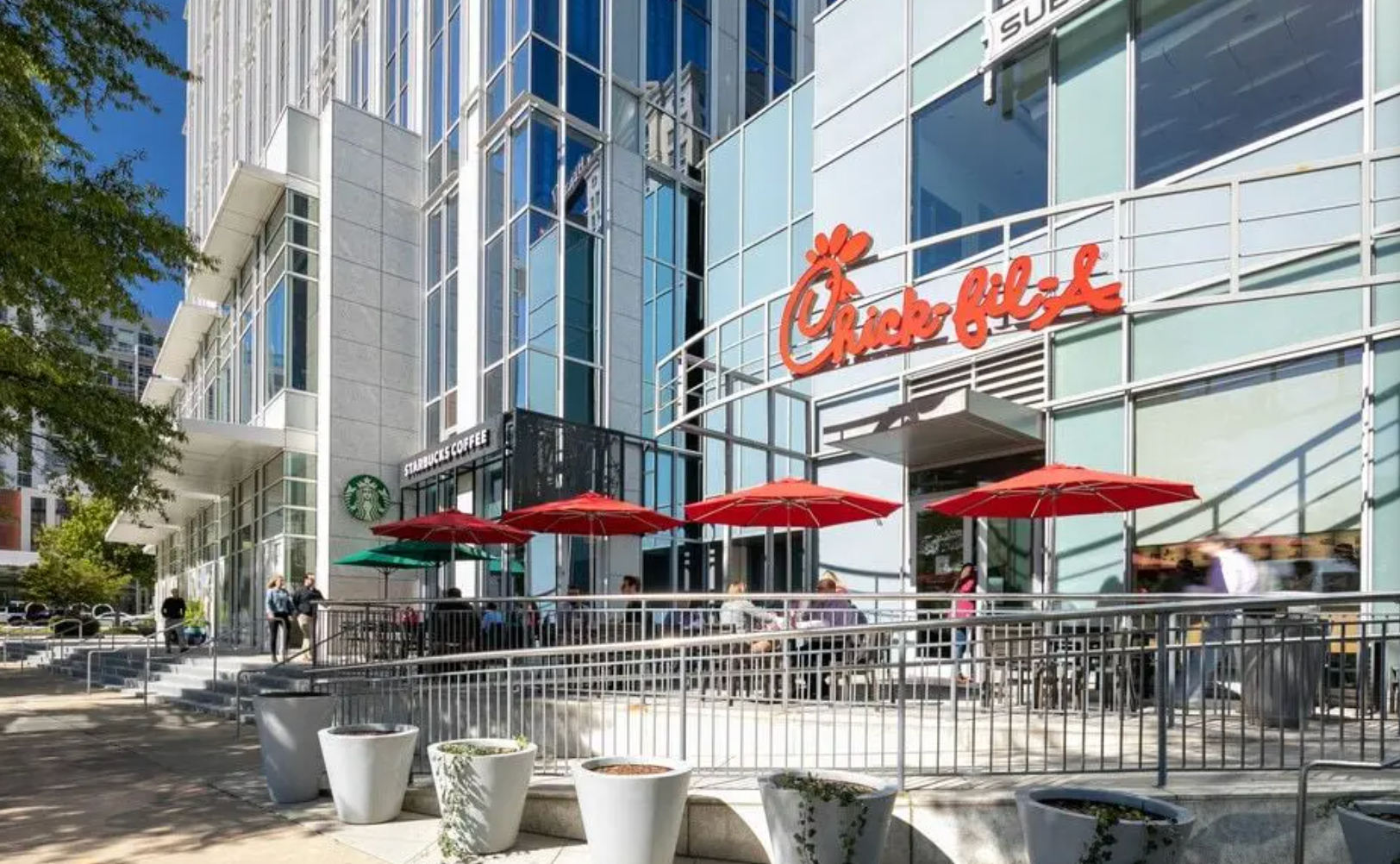 A Chick-fil-A restaurant entrance with outdoor seating, red umbrellas, and a modern glass office building background.