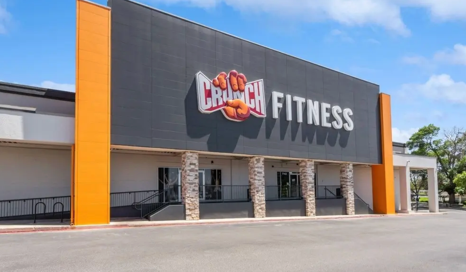 Exterior view of a Crunch Fitness gym building featuring a dark gray wall with orange pillars under a blue sky.