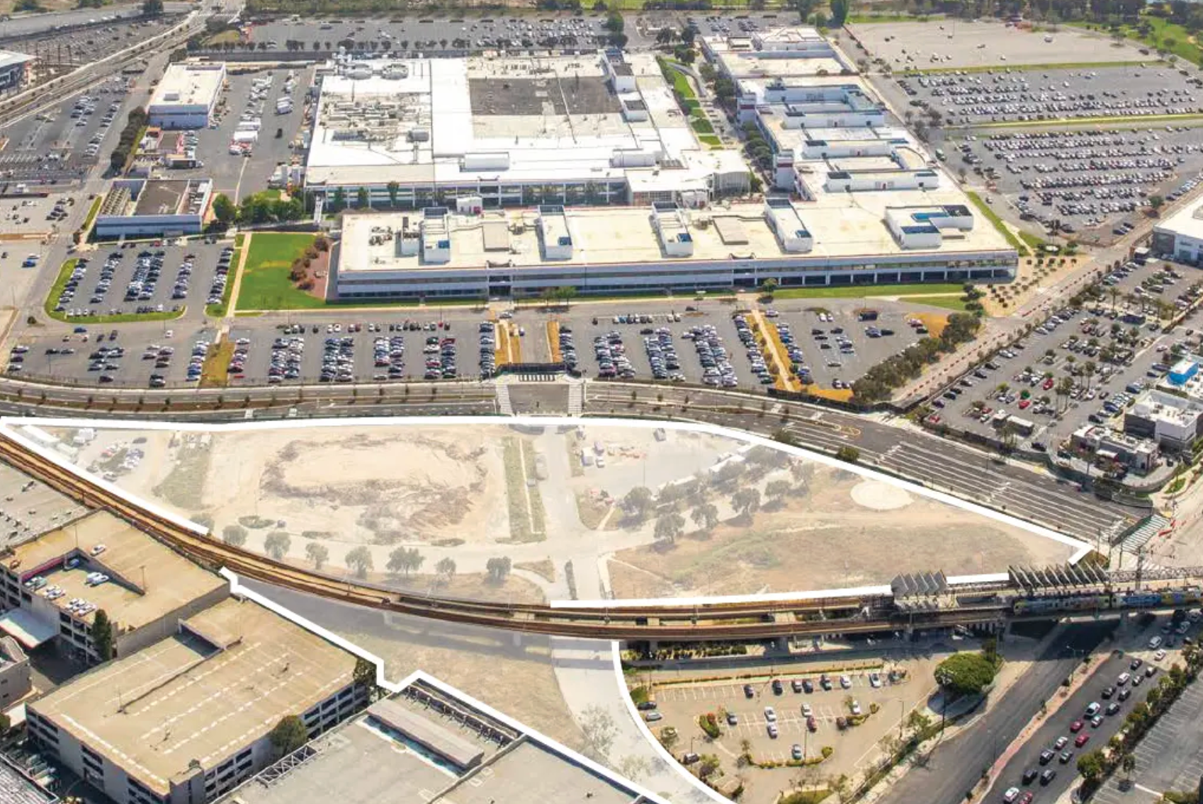 Aerial view of a sprawling commercial complex and parking lots bordering an expansive, undeveloped dirt lot and train tracks.