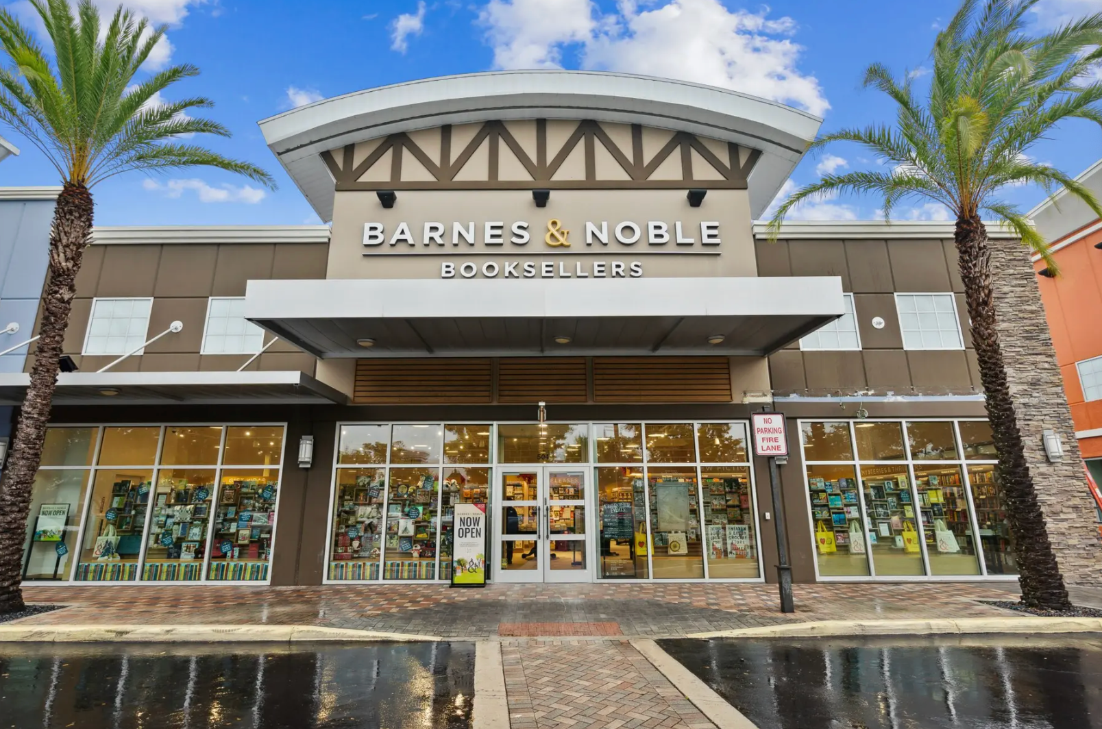 Exterior of a Barnes & Noble bookstore with large windows, a cream-colored facade, and palm trees under a blue sky.