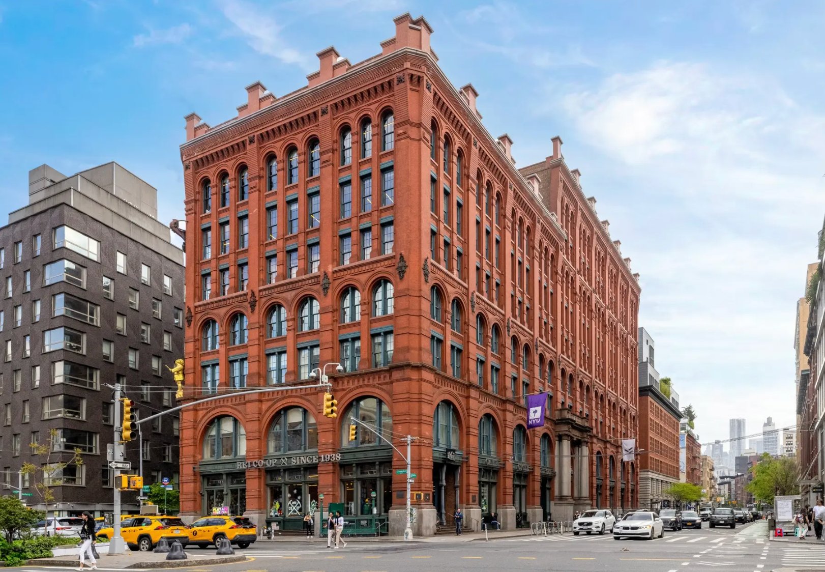 A multi-story, red brick historical building with arched ground-floor windows on a sunny, urban street corner.