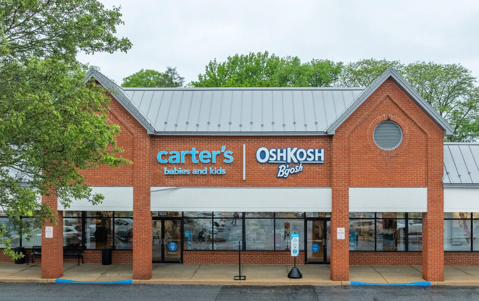 A brick storefront with signage for Carter’s and OshKosh B’gosh, featuring glass entry doors under a metal roof.