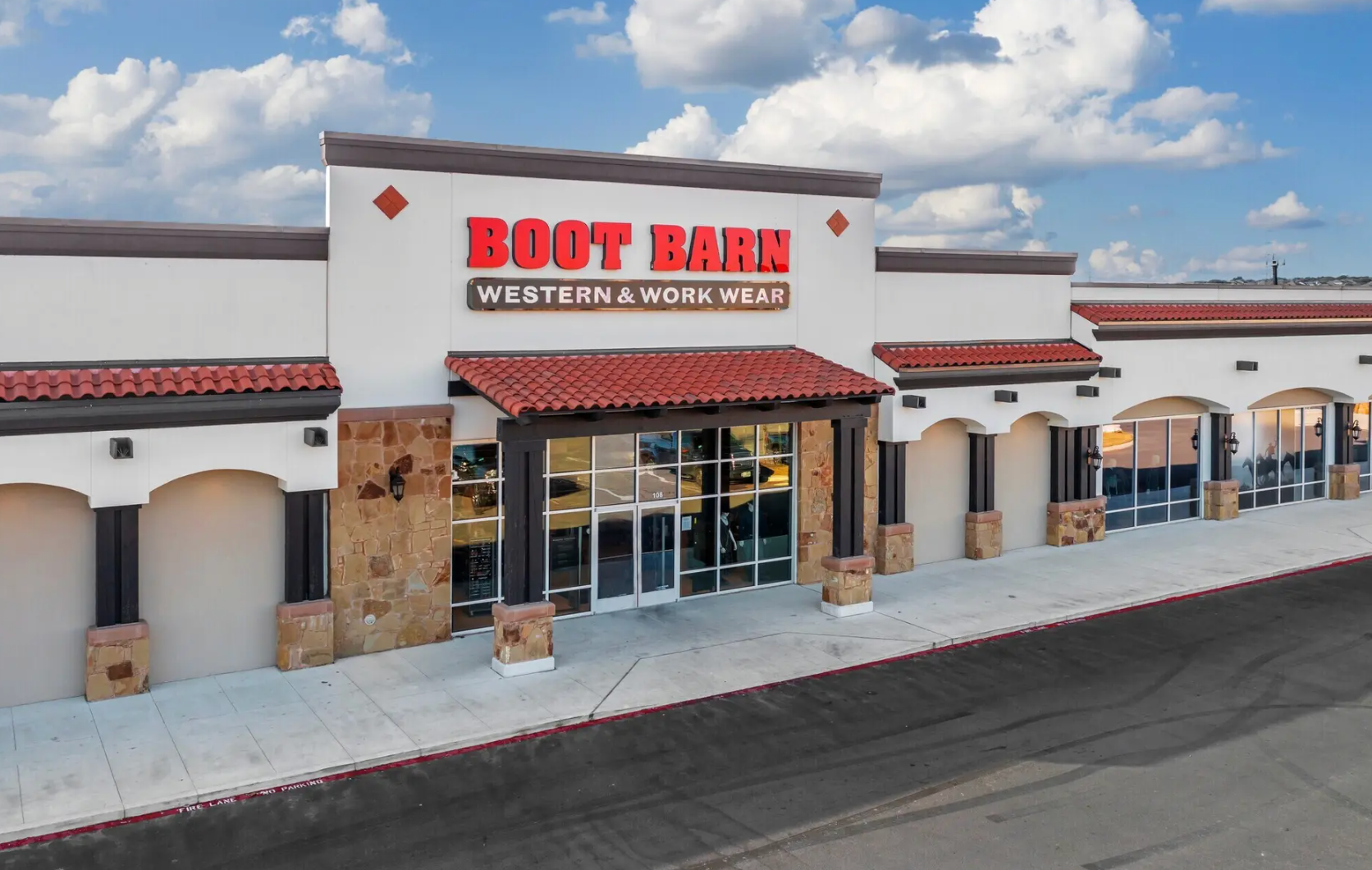 An exterior view of a Boot Barn retail store in a strip mall with white stucco walls, stone accents, and red roof tiles.