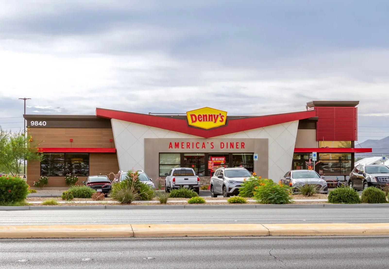 A modern Denny's restaurant exterior with a red roof, white accents, and cars parked in front under a cloudy sky.