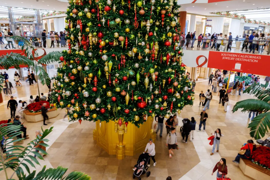 Shoppers walk past a large, decorated Christmas tree in a brightly lit, multistory shopping mall.