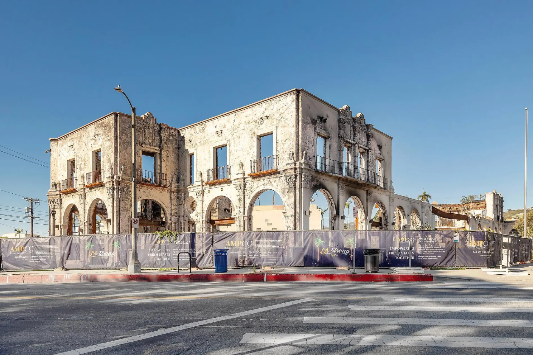 A partially demolished stone building with archways, surrounded by temporary construction fencing under a clear blue sky.