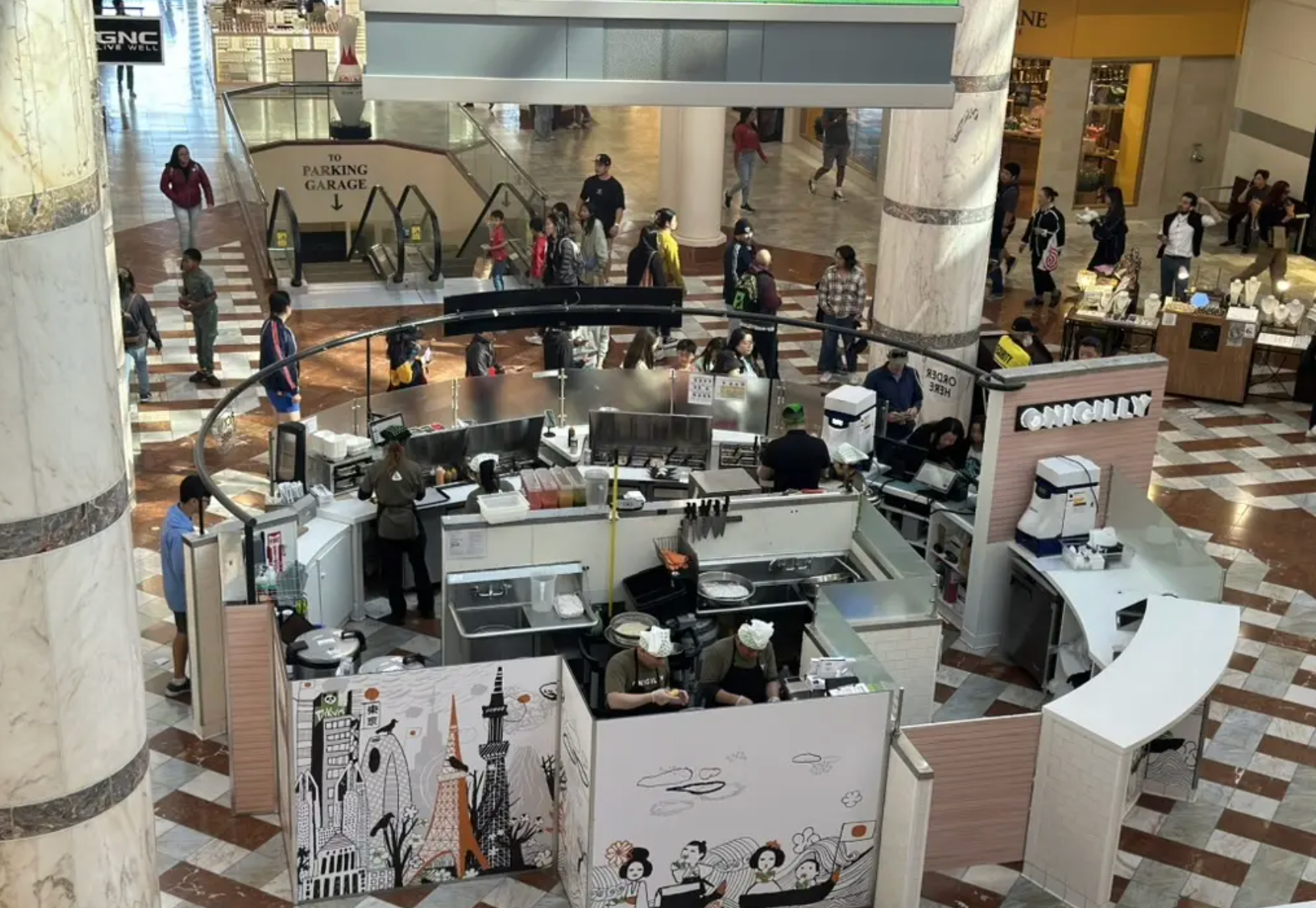 An elevated view of a central food kiosk inside a mall with patrons walking nearby and staff working behind the counter.