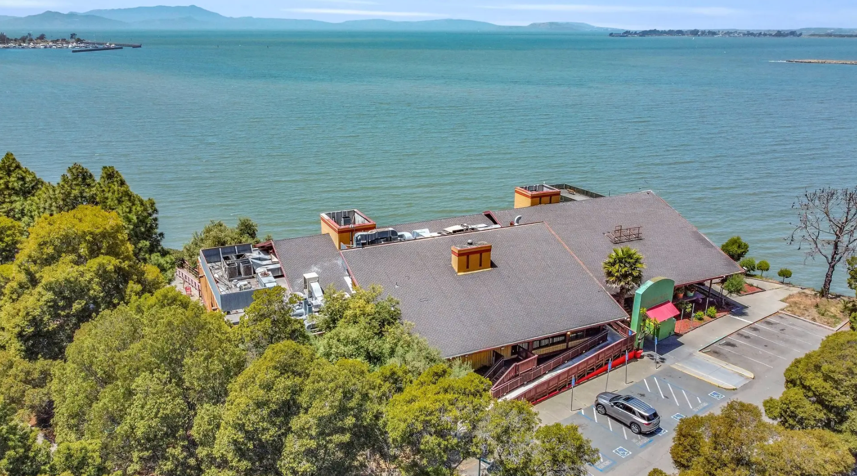 A high-angle view of a large building with a brown roof, nestled among lush trees beside a vast, blue body of water.