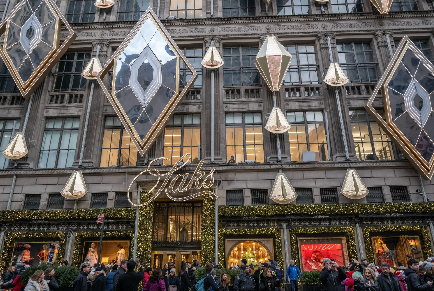 The Saks Fifth Avenue store exterior in New York, adorned with diamond-shaped lights and holiday greenery, with a crowd.