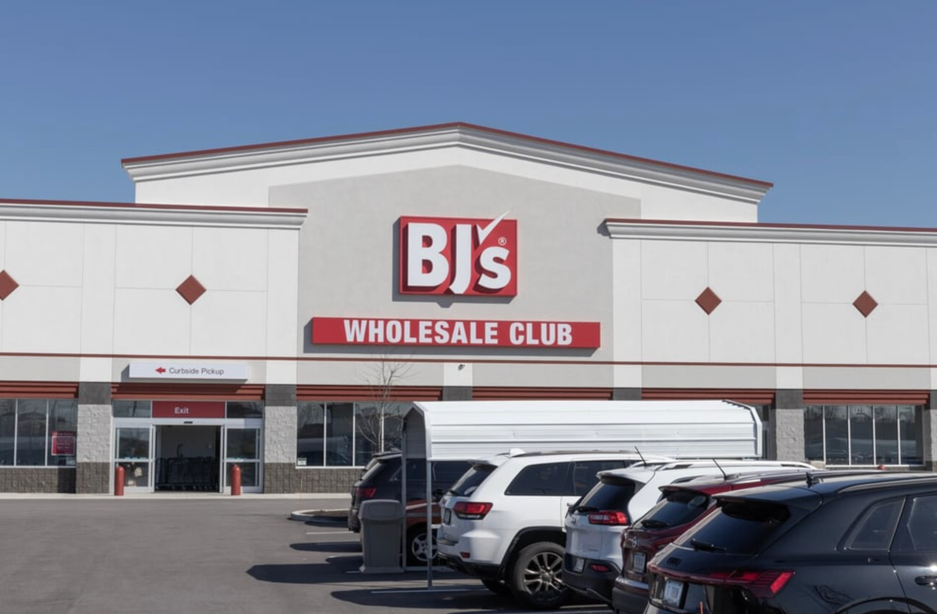 A BJ's Wholesale Club storefront under a clear blue sky, with a parking lot featuring several cars in the foreground.