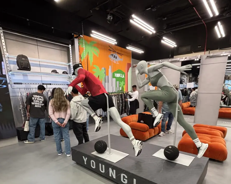 Two mannequins in athletic poses on a platform in a clothing store with shoppers browsing racks in the background.