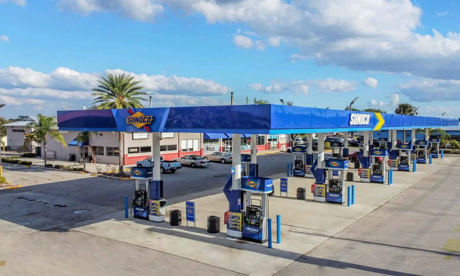 A row of fuel pumps under a long blue canopy at a gas station on a sunny day.