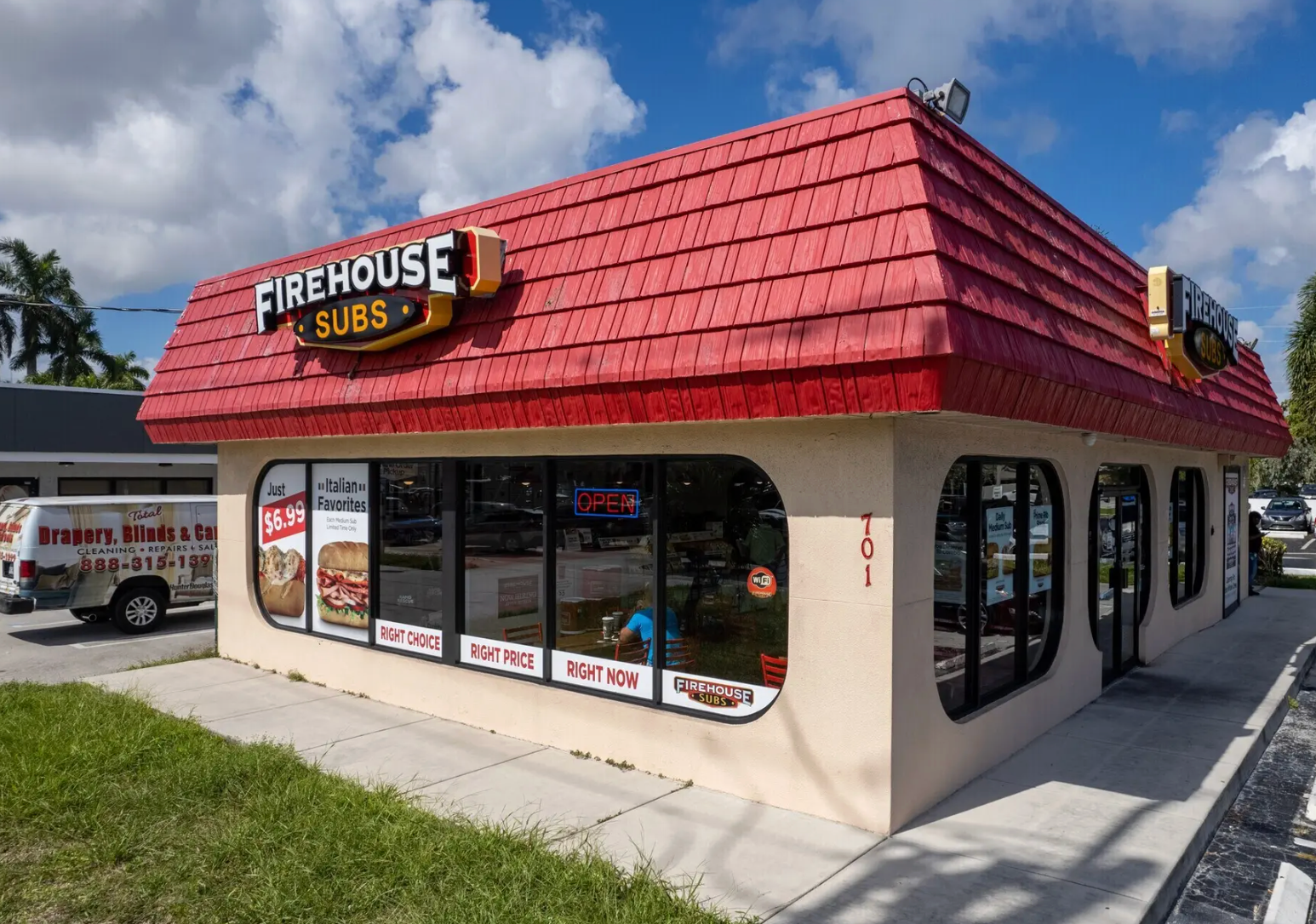 A corner view of a Firehouse Subs restaurant with a tan exterior, red tiled roof, and large glass windows.
