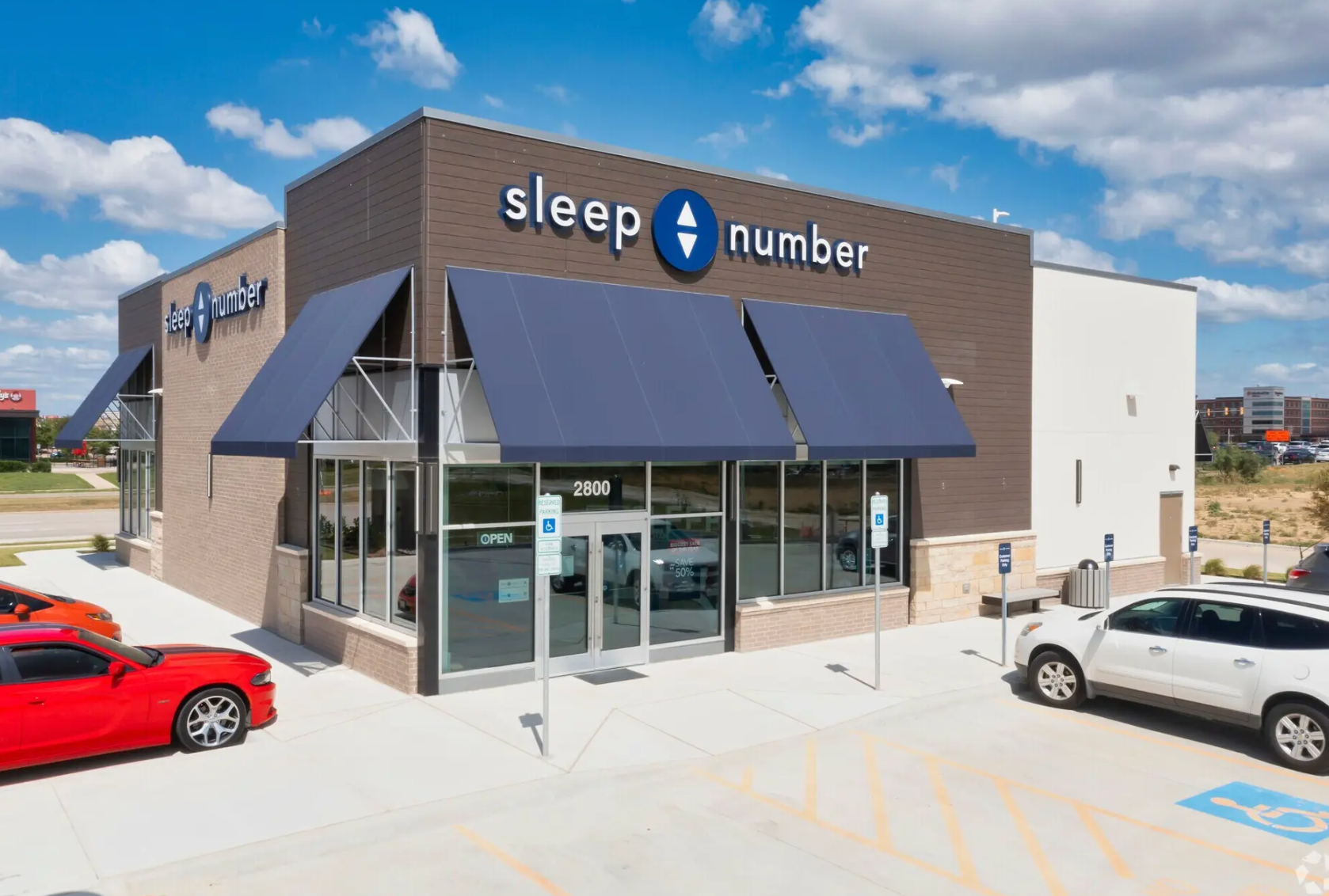 A Sleep Number retail store with a dark facade, blue awnings, and parking spots, including accessible spaces, out front.