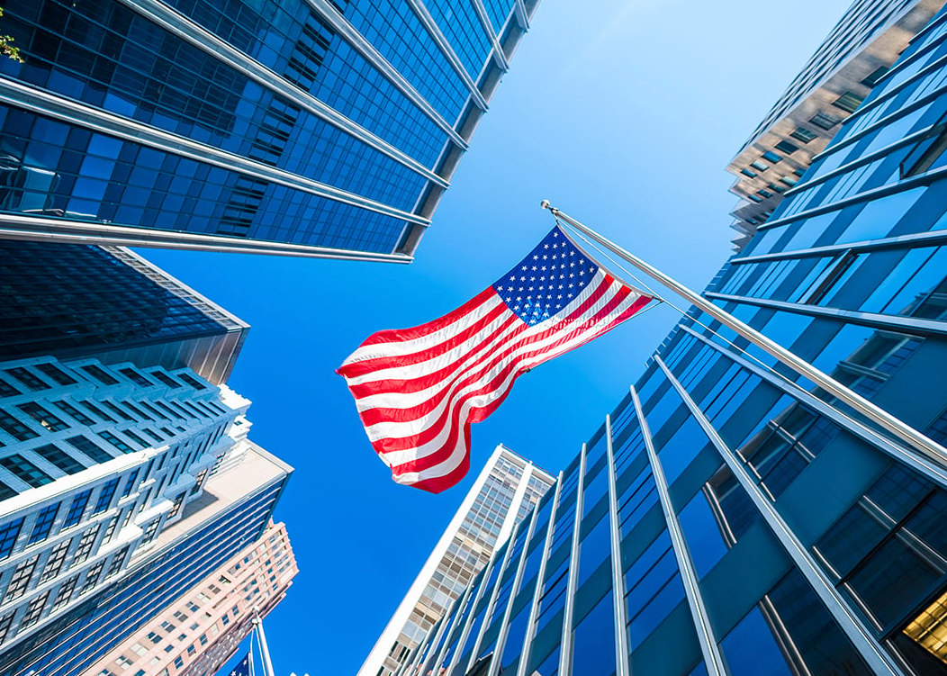 The American flag waves against a bright blue sky between towering glass skyscrapers, viewed from a low angle.