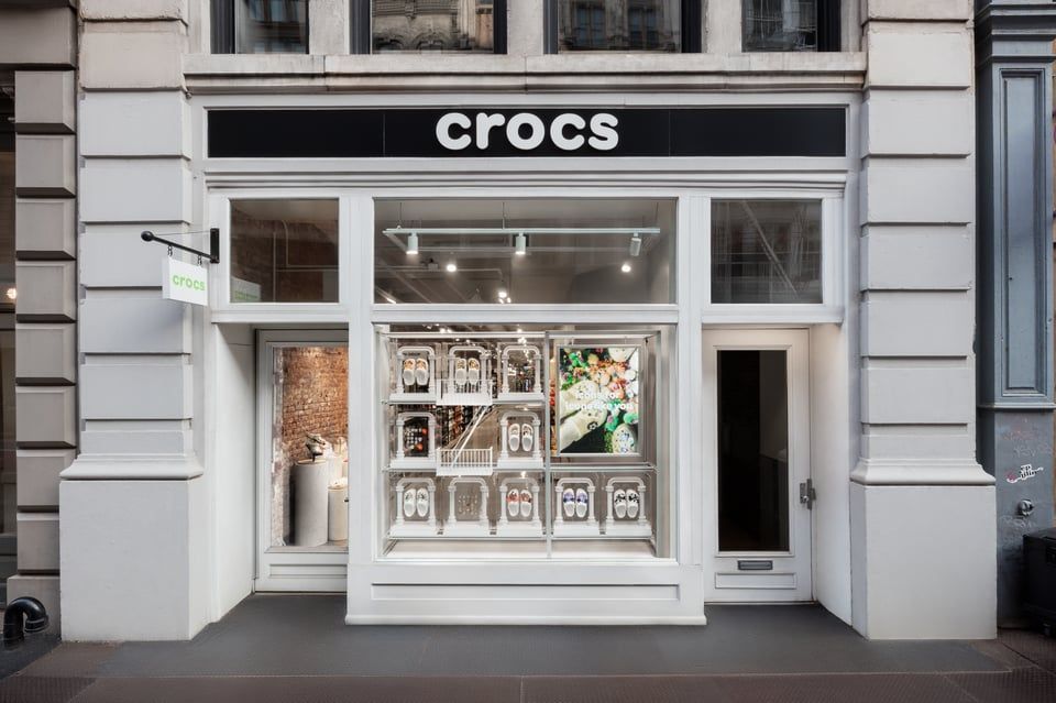 A storefront for a Crocs retail shop with a black sign, white facade, and window displays showing shoes and graphics.