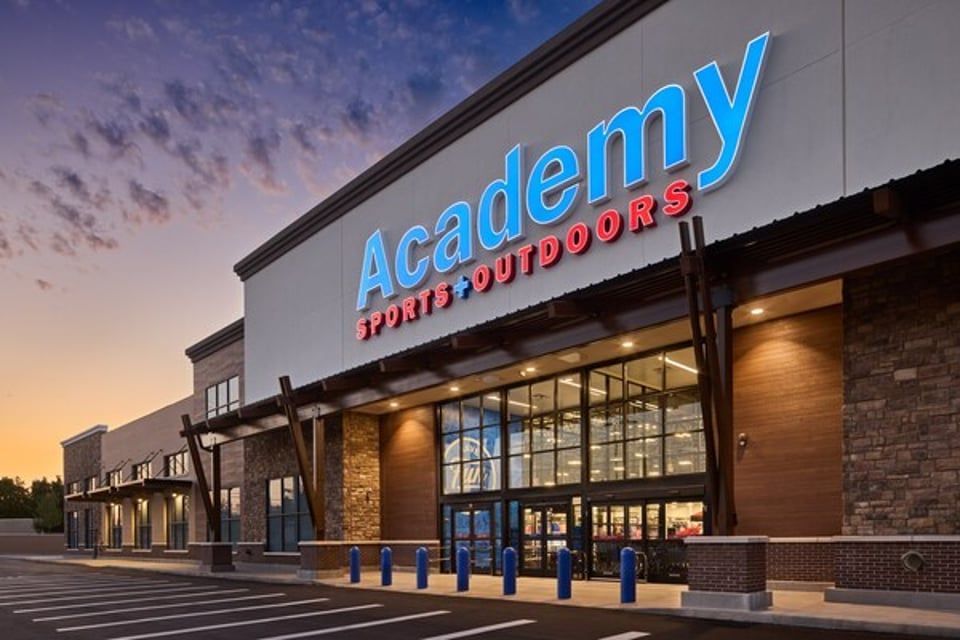 Academy Sports + Outdoors store exterior with stone facade, glass entrance, and parking lot under a sunset sky.