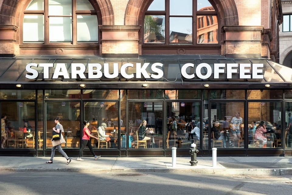 A storefront of a Starbucks Coffee cafe with large windows showing people inside and pedestrians walking on the sidewalk.