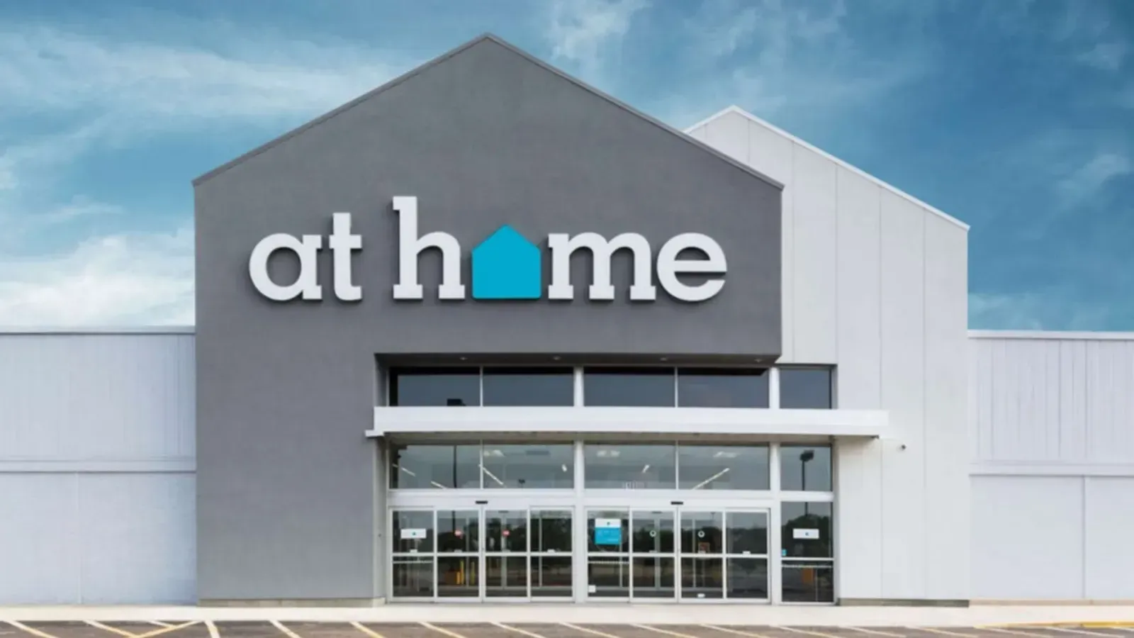 The entrance to an At Home store with a gray facade, white lettering, and a blue house icon over a glass-door storefront.