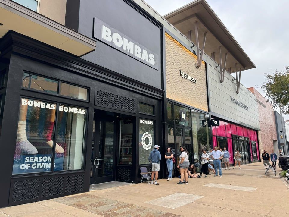 A storefront for Bombas with people standing on the sidewalk in front of it and adjacent shops under a cloudy sky.