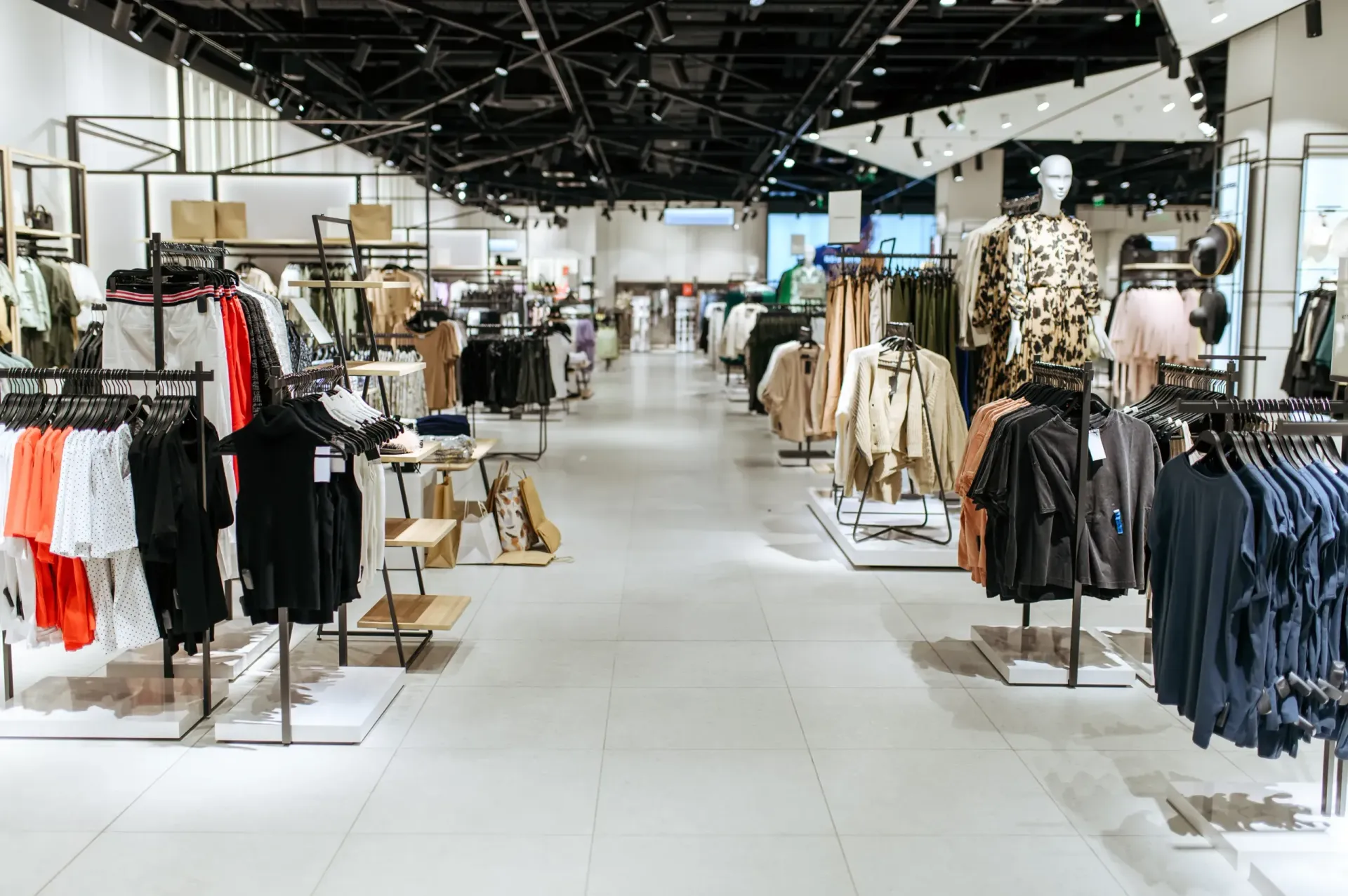A clothing store interior with rows of organized garment racks, shelves, and a bright tiled floor under a black ceiling.
